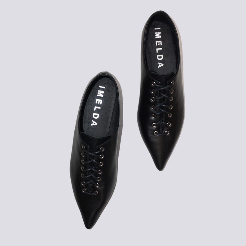 K&ouml;nnte beinhalten: Ein Paar schwarze Lederschn&uuml;rschuhe mit spitz zulaufenden Zehen. Die Schuhe tragen den Schriftzug "IMELDA" auf der Innenseite jedes Schuhs.