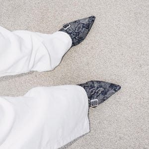 May include: A pair of black and white snake print shoes with a buckle closure. The shoes are worn with white pants.