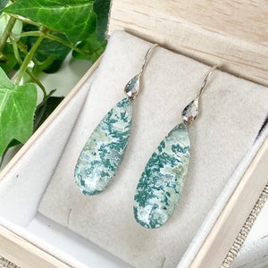 May include: A pair of silver teardrop earrings with green and white agate stones. The earrings are in a small wooden box with a white velvet lining.