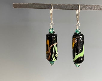 May include: A pair of black glass earrings with a swirl design in yellow and green. The earrings have a silver leverback closure and are accented with green beads. The earrings are cylindrical in shape.