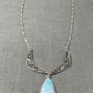 May include: A silver necklace with a teardrop-shaped blue opal pendant and a delicate silver chain. The pendant is surrounded by a silver filigree design.