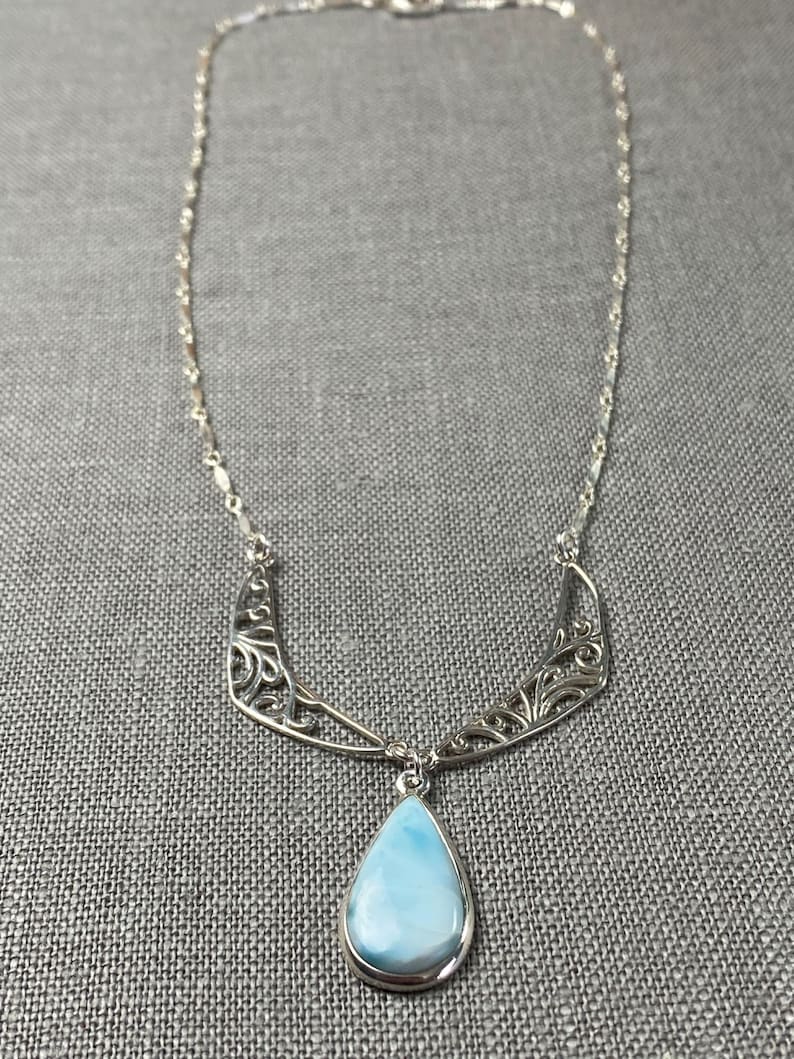 May include: A silver necklace with a teardrop-shaped blue opal pendant and a delicate silver chain. The pendant is surrounded by a silver filigree design.