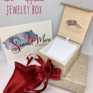 May include: A red satin jewelry pouch and a wooden jewelry box with a beige linen exterior. The box is open and has a white velvet interior. The box has a logo on the lid that says "Scarlet Mare Studio".