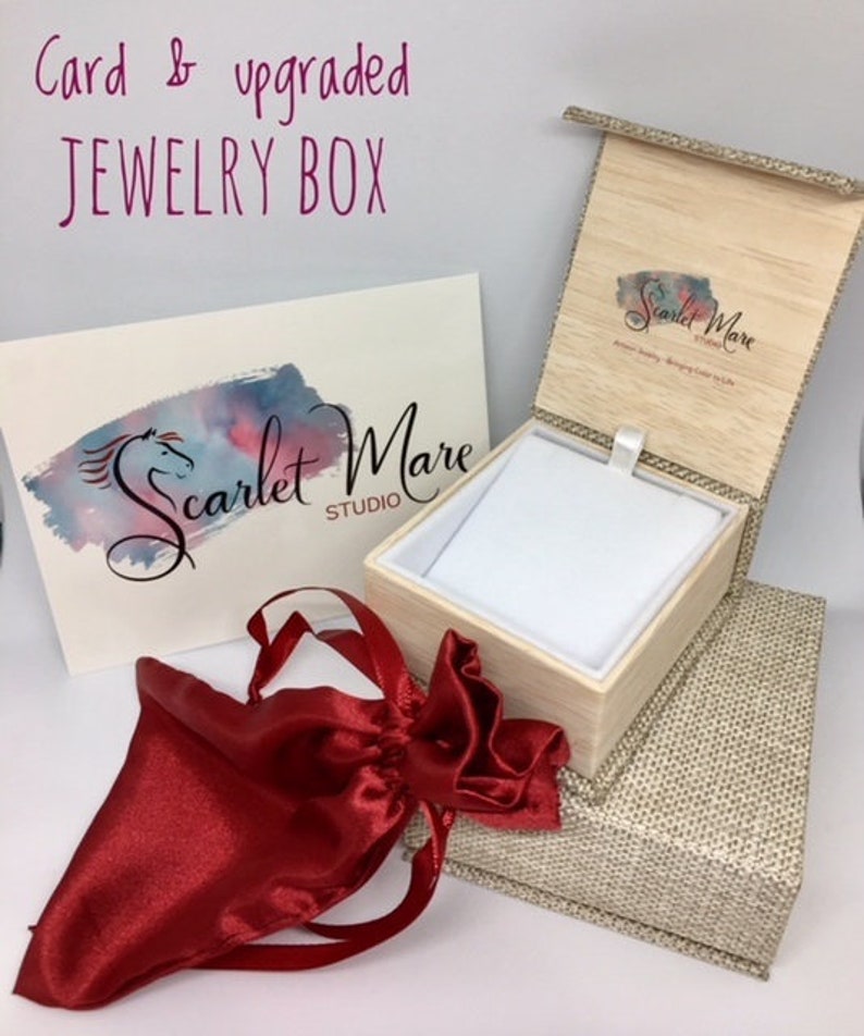 May include: A red satin jewelry pouch and a wooden jewelry box with a beige linen exterior. The box is open and has a white velvet interior. The box has a logo on the lid that says "Scarlet Mare Studio".