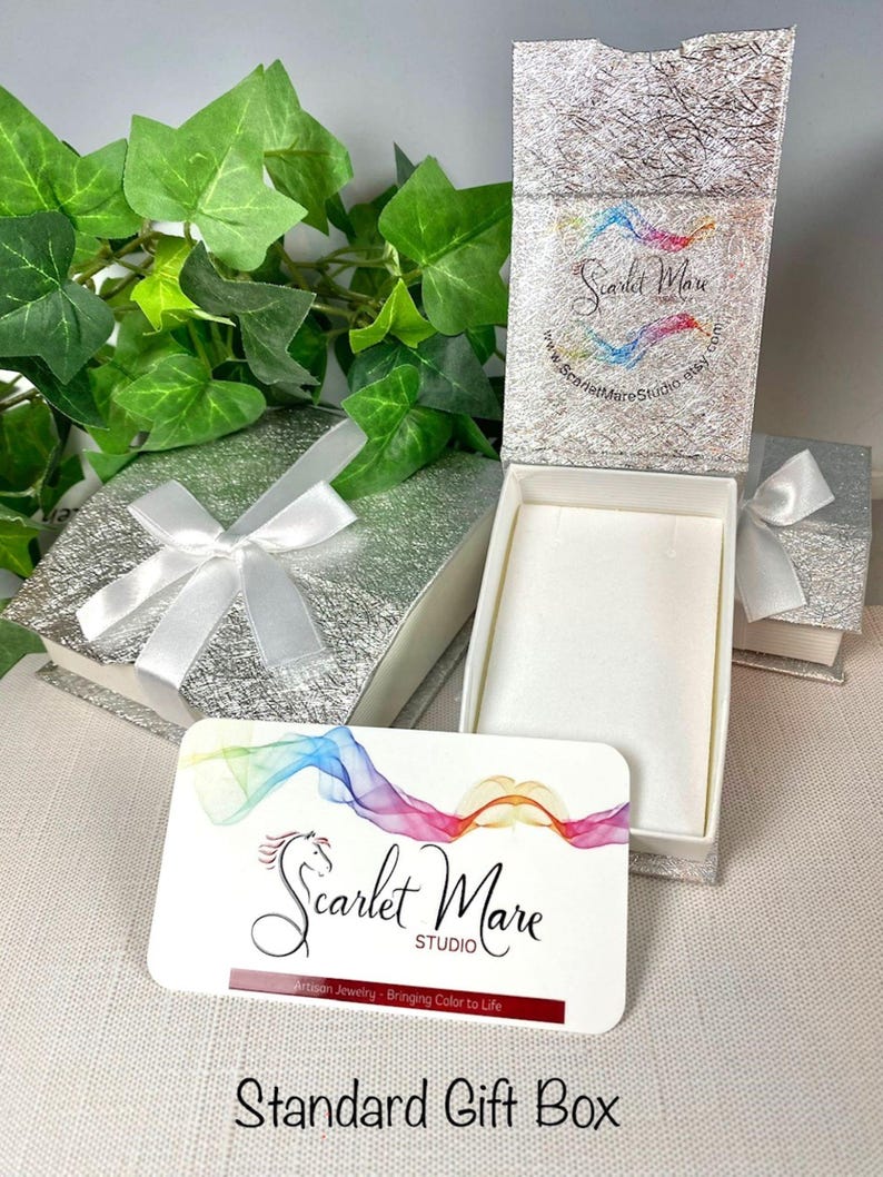 K&ouml;nnte beinhalten: Geschenkbox-Set mit silberner strukturierter Oberfl&auml;che, wei&szlig;em Band und wei&szlig;em Innenraum. Eine Visitenkarte mit dem Text "Scarlet Mare Studio" und "Standard Gift Box" ist ebenfalls sichtbar. Gr&uuml;nes Efeu ist im Hintergrund.