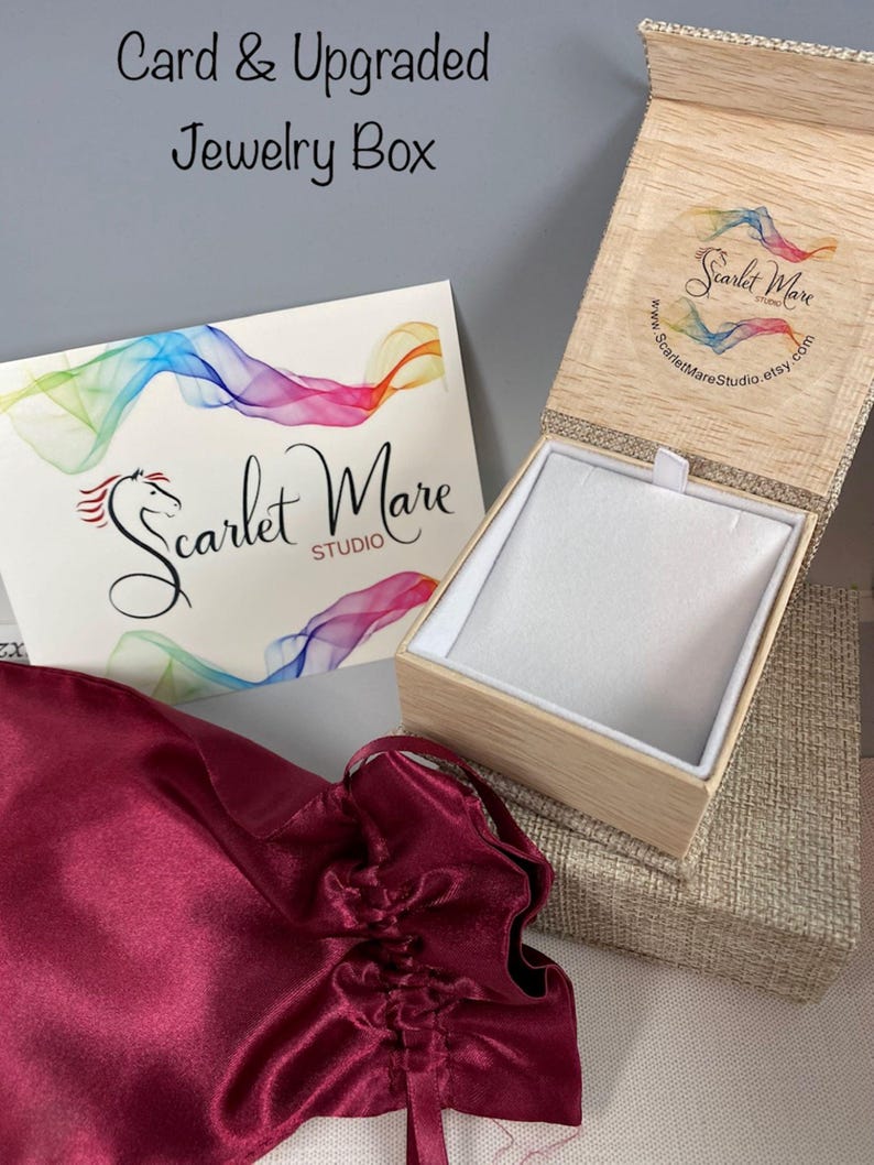 K&ouml;nnte beinhalten: Eine offene Holzschmuckschatulle mit wei&szlig;em Innenraum, eine Karte mit farbenfrohen abstrakten Designs und ein burgunderfarbener Satinbeutel. Die Karte und die Schachtel tragen den Text "Scarlet Mare Studio". Der Text "Card & Upgraded Jewelry Box" ist ebenfalls sichtbar.