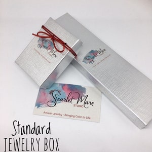 May include: A silver jewelry box with a red ribbon and the text "Scarlet Mare Studio" printed on the box. The text "Artisan Jewelry - Bringing Color to Life" is also printed on the box. The box is labeled "Standard Jewelry Box".