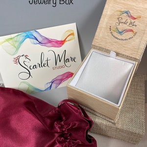 May include: A burgundy satin jewelry pouch with a drawstring closure, a wooden jewelry box with a linen exterior, and a card with the text "Scarlet Mare Studio" and a rainbow design.