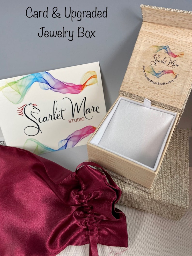 May include: A burgundy satin jewelry pouch with a drawstring closure, a wooden jewelry box with a linen exterior, and a card with the text "Scarlet Mare Studio" and a rainbow design.
