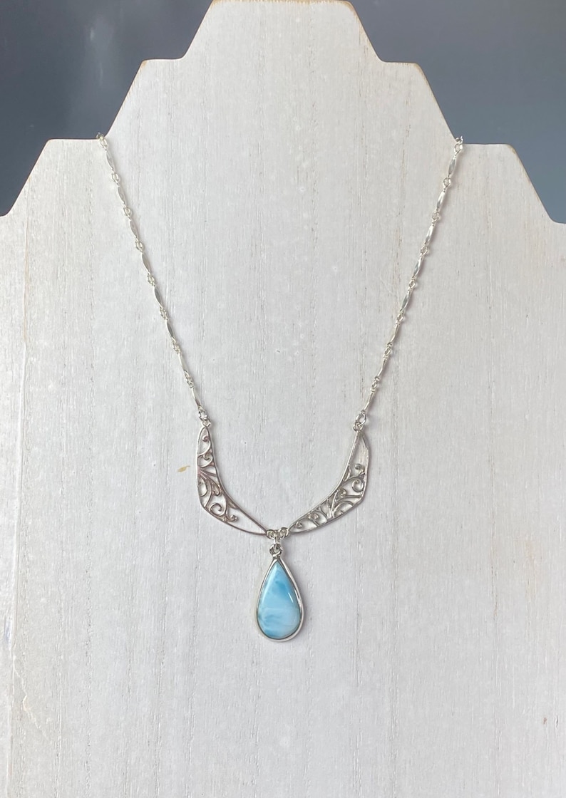 May include: A silver necklace with a teardrop-shaped blue gemstone pendant. The necklace features a delicate, filigree design.