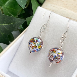 Könnte beinhalten: Ein Paar silberne Ohrringe mit einem runden, mehrfarbigen Glasperlen an jedem Ohrring. Die Perlen sind mit kleinen Glasstücken in den Farben Rot, Blau, Grün, Gelb und Silber gefüllt.