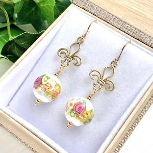 May include: A pair of gold earrings with a white glass bead featuring a pink floral design and a gold fleur-de-lis charm. The earrings are displayed in a small wooden box with a white velvet lining.