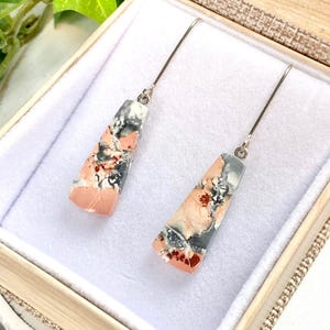 Könnte beinhalten: Ein Paar silberne Ohrringe mit einem Anhänger aus rosa, grauem und weißem Stein. Die Ohrringe befinden sich in einer kleinen Schachtel.