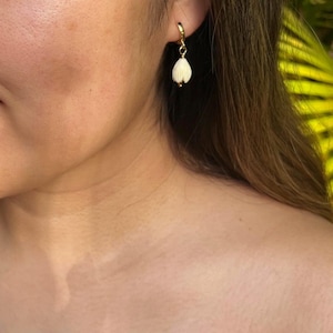 May include: A close-up of a gold hoop earring with a white, teardrop-shaped pendant. The pendant has a textured, layered appearance. The earring is worn on a person's ear, with the focus on the jewelry.