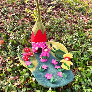 May include: A whimsical felt fairy garden scene featuring a red and green mushroom-shaped house, small felt fairies, and colorful flowers. The scene is set on a blue and green felt base, surrounded by greenery.