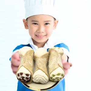 May include: A young person wearing a chef's hat and blue shirt holds a plate with three cannoli. The cannoli have tan, cream, and brown accents, with a variety of fillings. The plate has a gold rim.