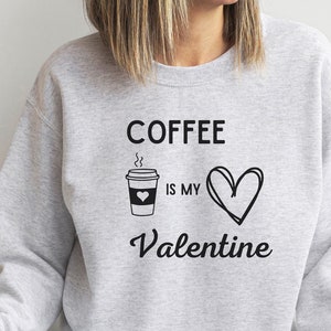 Op de afbeelding: Een grijs sweatshirt met de tekst "Coffee is my Valentine" en een tekening van een koffiebeker en een hart.