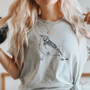 Könnte beinhalten: Ein graues T-Shirt mit einer schwarz-weißen Illustration eines Vogels, der auf einem Ast sitzt.
