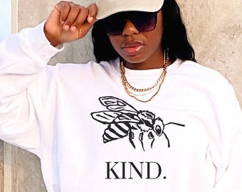 May include: White sweatshirt with a black bee graphic and the word "KIND." printed below. The model is wearing a beige baseball cap and black shorts.