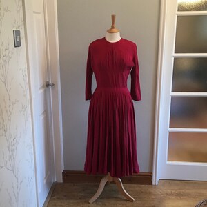 Può includere: Un abito rosso vintage con un corpetto aderente e una gonna ampia. L'abito è su un manichino e ha un design semplice ed elegante.