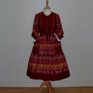 Könnte beinhalten: Ein rotes Kleid mit einem geometrischen Muster in Orange-, Gelb-, Blau- und Rosatönen. Das Kleid hat ein tailliertes Oberteil und einen weiten Rock mit Taschen.