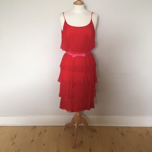 May include: A red tiered dress with a pink ribbon belt. The dress is sleeveless and has a ruffled neckline.