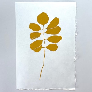 May include: A botanical art print featuring a mustard-yellow leaf design on a white background. The leaf has a long stem and several rounded leaves. The paper has a deckled edge, adding a handmade feel to the artwork.
