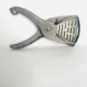 May include: A silver-colored potato ricer, a kitchen utensil used to mash cooked potatoes. The ricer has a handle and a perforated basket for pressing the potatoes through. The utensil is made of metal and has a textured surface.