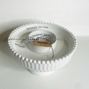 May include: White ceramic serving dish with a raised base and beaded rim. The dish has a central, smaller bowl and is tied with black and white twine. Text on the dish reads "Shrimply the best" and "Cocktail hour".