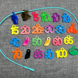 May include: A collection of colorful plastic charms featuring numbers from 5 to 100, along with symbols like a person, a bicycle, a swimmer, and a foot. These charms are arranged inside a light blue circular cord, each with a small heart-shaped loop.