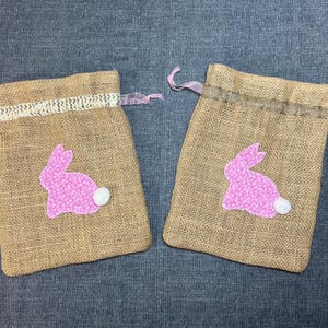 May include: Two burlap drawstring bags with pink bunny appliqués and white pom-pom tails. The bunnies are made of pink patterned fabric. The bags have a decorative trim and pink ribbon drawstrings. Perfect for Easter gifts.