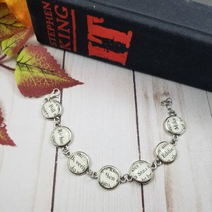 Op de afbeelding: Een zilveren armband met ronde bedels met tekst uit het boek "IT" van Stephen King. De bedels bevatten de namen van de personages: Bill, Richie, Beverly, Ben, Stan, Eddie en Mike.