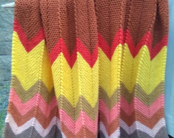 May include: A handmade, chevron-patterned blanket in warm tones of brown, red, yellow, and pink. The knitted blanket is displayed, showcasing its textured surface and vibrant color scheme. The zig-zag design adds a touch of retro style to this cozy home decor item.