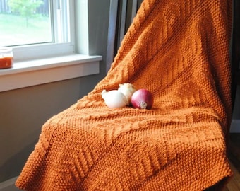 May include: An orange knitted blanket draped over a wooden chair. The blanket has a textured pattern and is folded to display its design. Two onions sit on top of the blanket near a window.