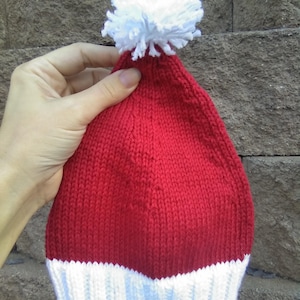 May include: A red knit hat with a white pom-pom on top and a white brim. The hat is held by a hand against a textured background.