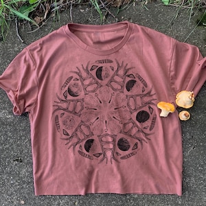 May include: A mauve cropped t-shirt with a black and white graphic print of a mandala design featuring antlers, moons, and feathers.