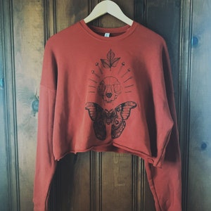 May include: A cropped, rust-colored sweatshirt with a black and white graphic print of a butterfly, skull, and leaf. The graphic features a skull with a leaf growing from the top and surrounded by pins.
