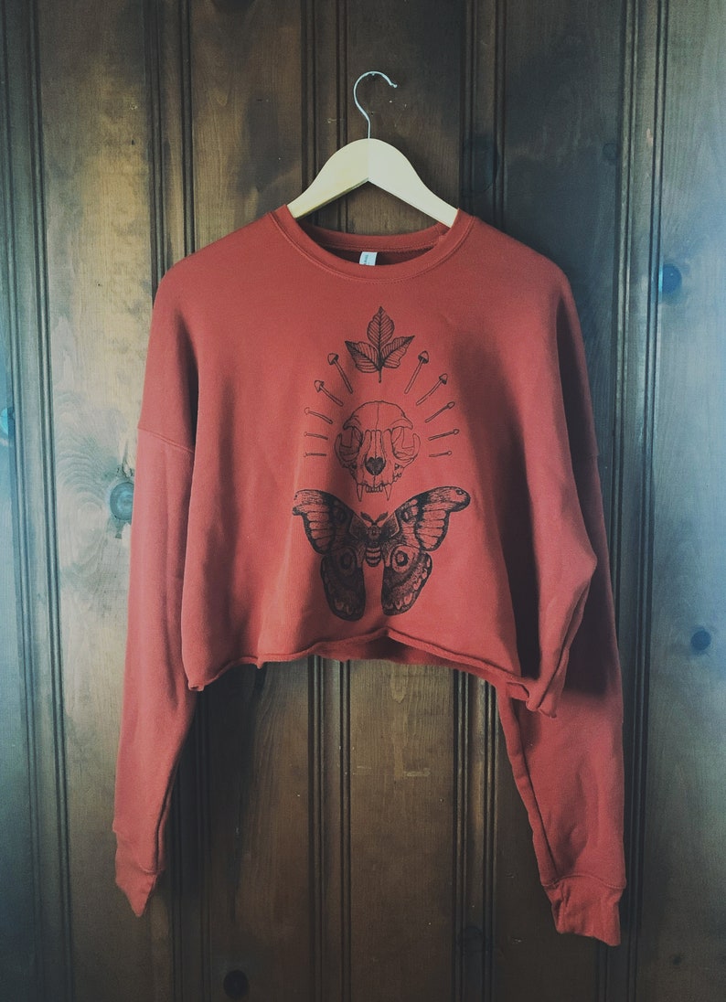 May include: A cropped, rust-colored sweatshirt with a black and white graphic print of a butterfly, skull, and leaf. The graphic features a skull with a leaf growing from the top and surrounded by pins.