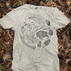 May include: A light gray t-shirt with a black and white illustration of a toad, mushrooms, leaves, a flower, a butterfly, and an antler. The illustration is in a circle on the front of the shirt.