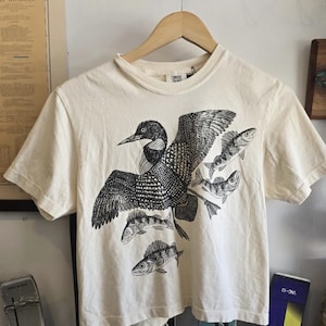 May include: Off-white t-shirt with a black and white graphic of a loon and fish. The bird has its wings spread, and the design is printed on a short-sleeved tee. The shirt is displayed on a wooden hanger.