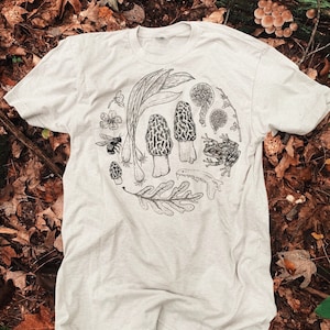 May include: A light grey t-shirt with a black and white illustration of a mushroom, a frog, a bee, and other forest elements. The illustration is in a circle on the front of the shirt.