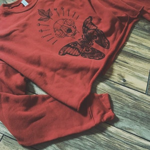 May include: A red sweatshirt with a black graphic print of a moth, skull, and leaves. The graphic is centered on the front of the sweatshirt.