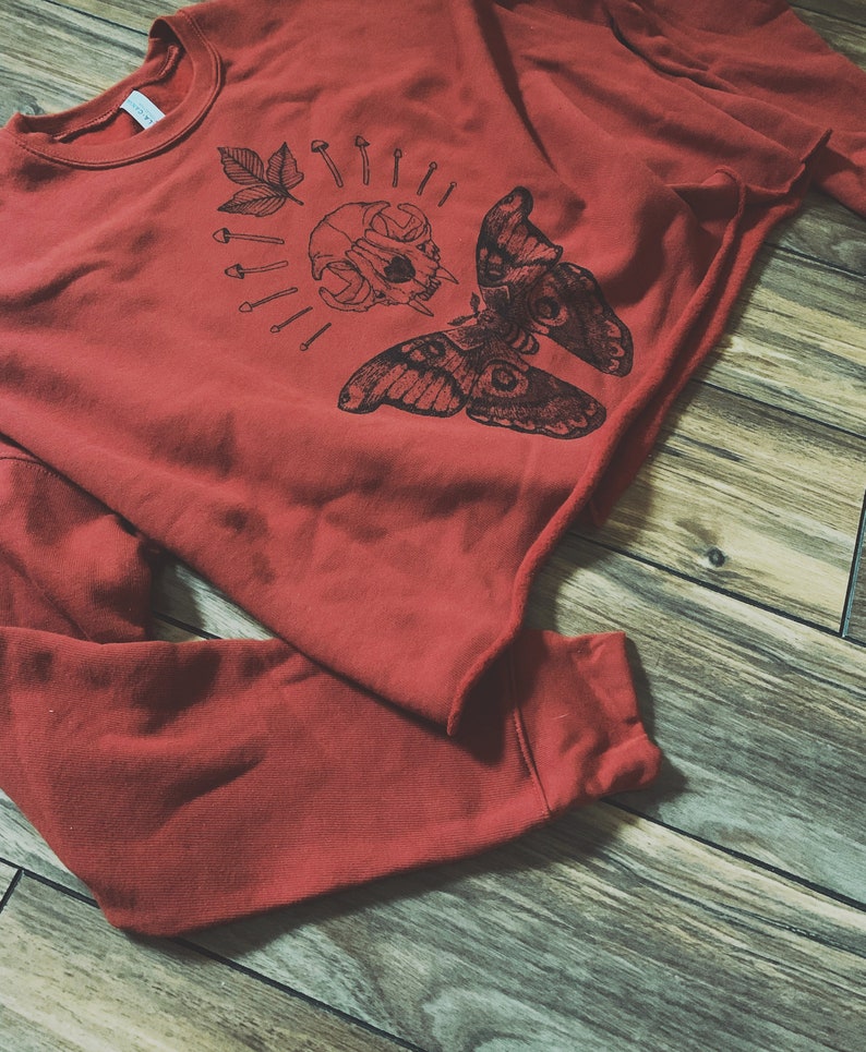 May include: A red sweatshirt with a black graphic print of a moth, skull, and leaves. The graphic is centered on the front of the sweatshirt.