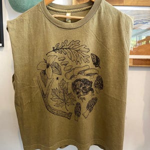 May include: A sleeveless, olive green t-shirt featuring a black and white illustration of a frog, mushrooms, leaves, and a deer antler. The shirt has a round neck and a relaxed fit.