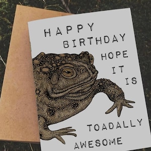 Könnte beinhalten: Eine Grußkarte mit einer Illustration einer braunen Kröte und dem Text "HAPPY BIRTHDAY HOPE IT IS TOADALLY AWESOME". Die Karte ist hellgrau mit schwarzem Text und einer braunen Kröte. Ein brauner Umschlag befindet sich hinter der Karte.