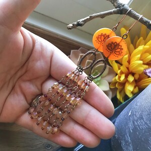 May include: Copper-colored dangle earrings with orange circular accents featuring a lotus flower design. The earrings have multiple strands of small, faceted beads in shades of amber and red, creating a cascading effect. The earrings are held in a hand.