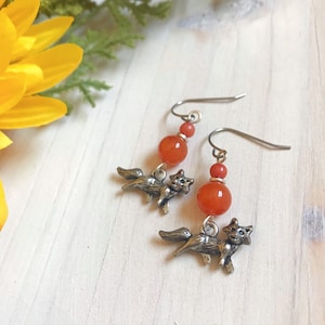 Puede incluir: Pendientes colgantes con pequeños dijes de zorro plateados y cuentas de piedras preciosas de color naranja rojizo. Los pendientes tienen ganchos plateados. Se muestran sobre una superficie de madera blanca con flores amarillas.