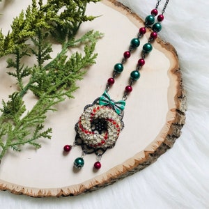 Pode incluir: Um colar festivo com um pingente de coroa adornado com cristais vermelhos e transparentes. O pingente é adornado com um laço verde e é suspenso de uma corrente com contas verdes e vermelhas alternadas. O colar é exibido em uma fatia de madeira.