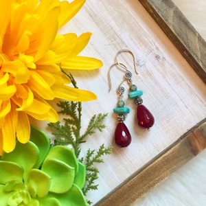Può includere: Un paio di orecchini pendenti in argento con un design a spirale. Ogni orecchino presenta una pietra rosso scuro sfaccettata a forma di goccia, perline turchesi e una perlina d'argento. Gli orecchini sono esposti su un vassoio di legno con fiori gialli e succulente verdi.