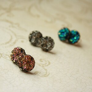 May include: Close-up of three pairs of druzy stud earrings in various colors. One pair is a mix of pink and gold, another is a dark gray, and the last is a vibrant teal. The earrings have a textured, sparkling appearance.